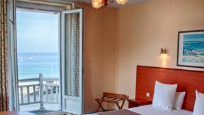 Een hotelkamer met een bed, bureau, stoel, balkon en een uitzicht op zee, plus een kroonluchter.