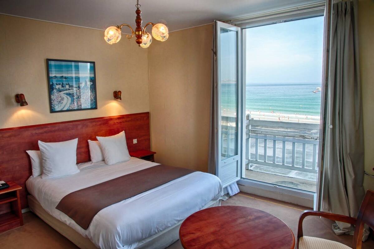 Une chambre d’hôtel avec un grand lit, une tête de lit en bois, un tableau au mur, un lustre et un balcon avec vue sur la plage.