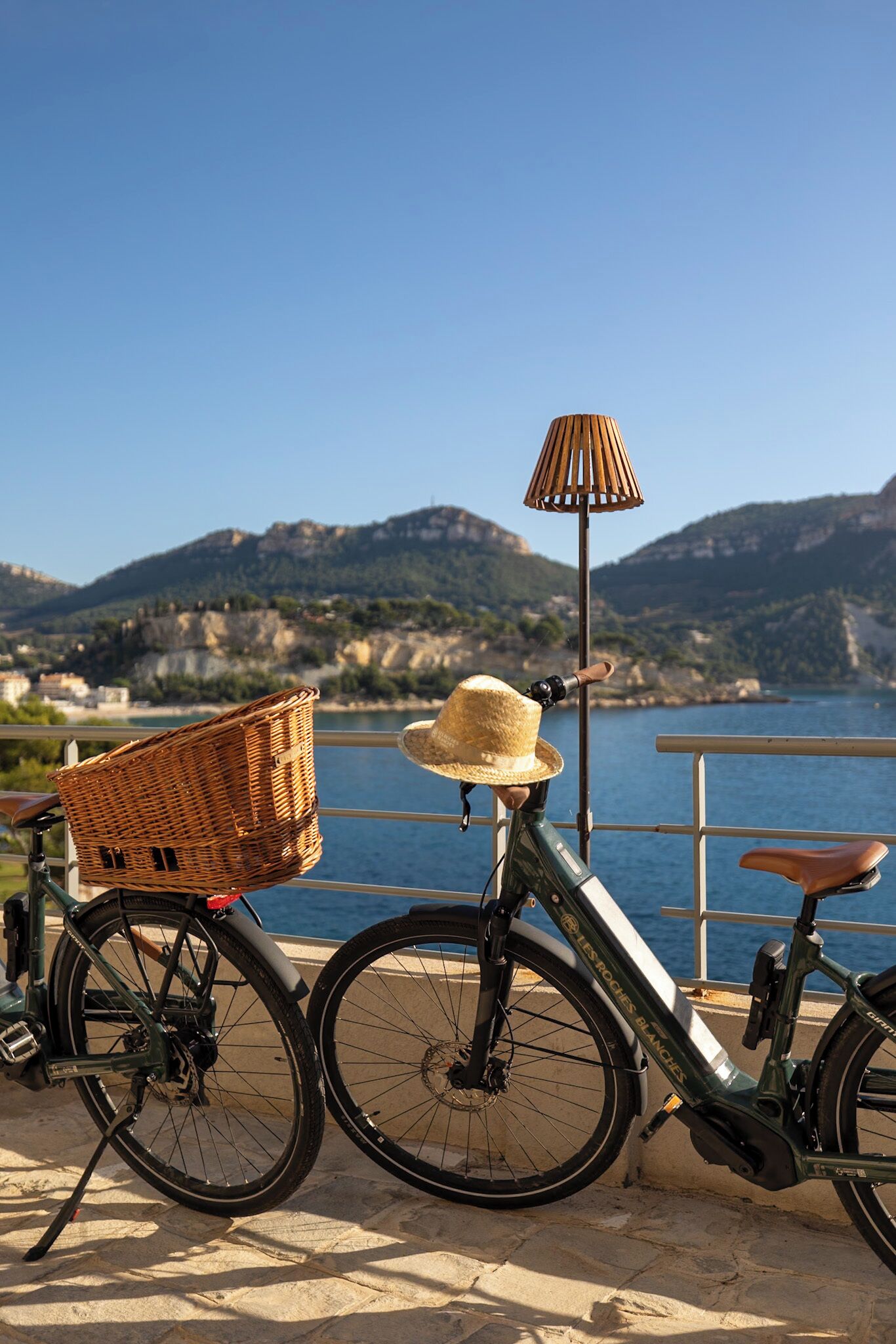 Una bicicleta con una cesta y un sombrero de sol apoyada en una barandilla junto al agua, con montañas al fondo.