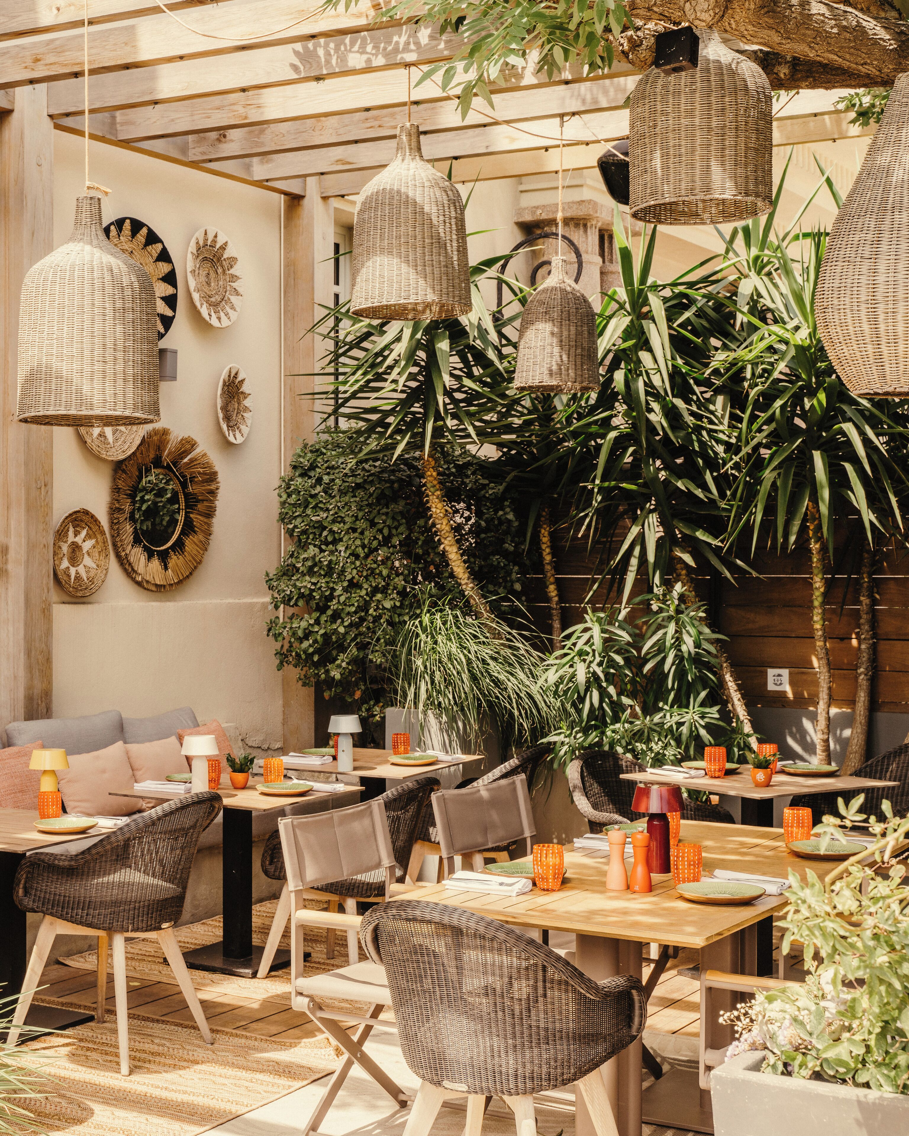Un área de comedor al aire libre con mobiliario de mimbre, lámparas colgantes de mimbre y diversas plantas.