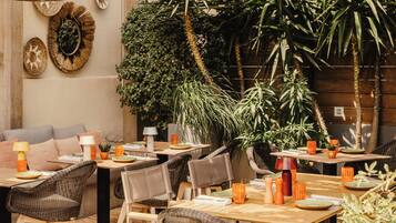 Un área de comedor al aire libre con mobiliario de mimbre, lámparas colgantes de mimbre y diversas plantas.