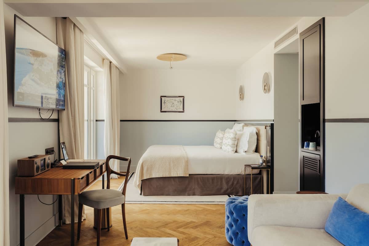 A modern hotel room with a wooden desk, a comfortable chair, a large bed, a sofa, and a television on the wall.