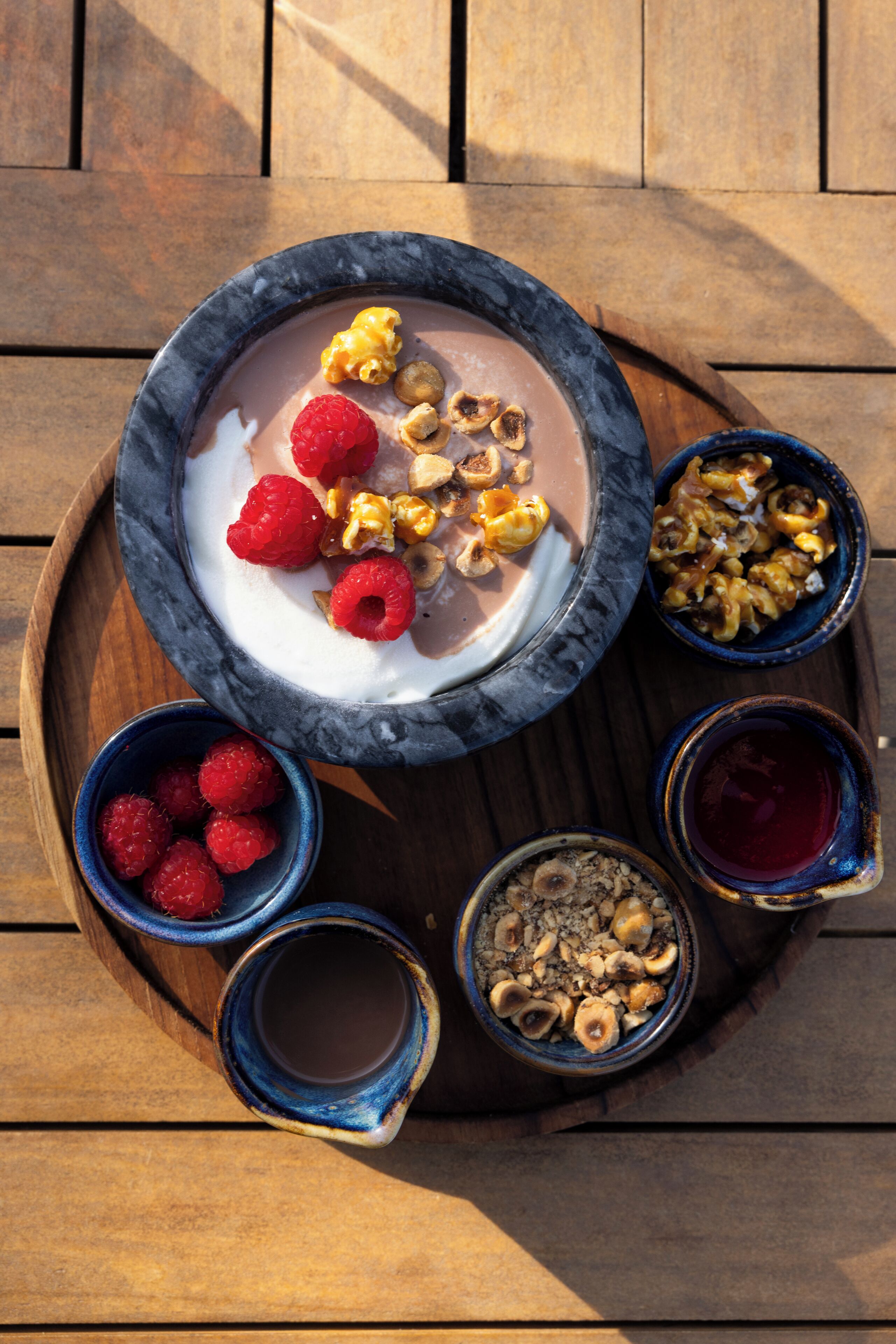 Una bandeja de postres con un tazón de yogur cubierto de frambuesas y nueces, acompañado de tazones de frambuesas, nueces y un pequeño tazón de salsa roja.