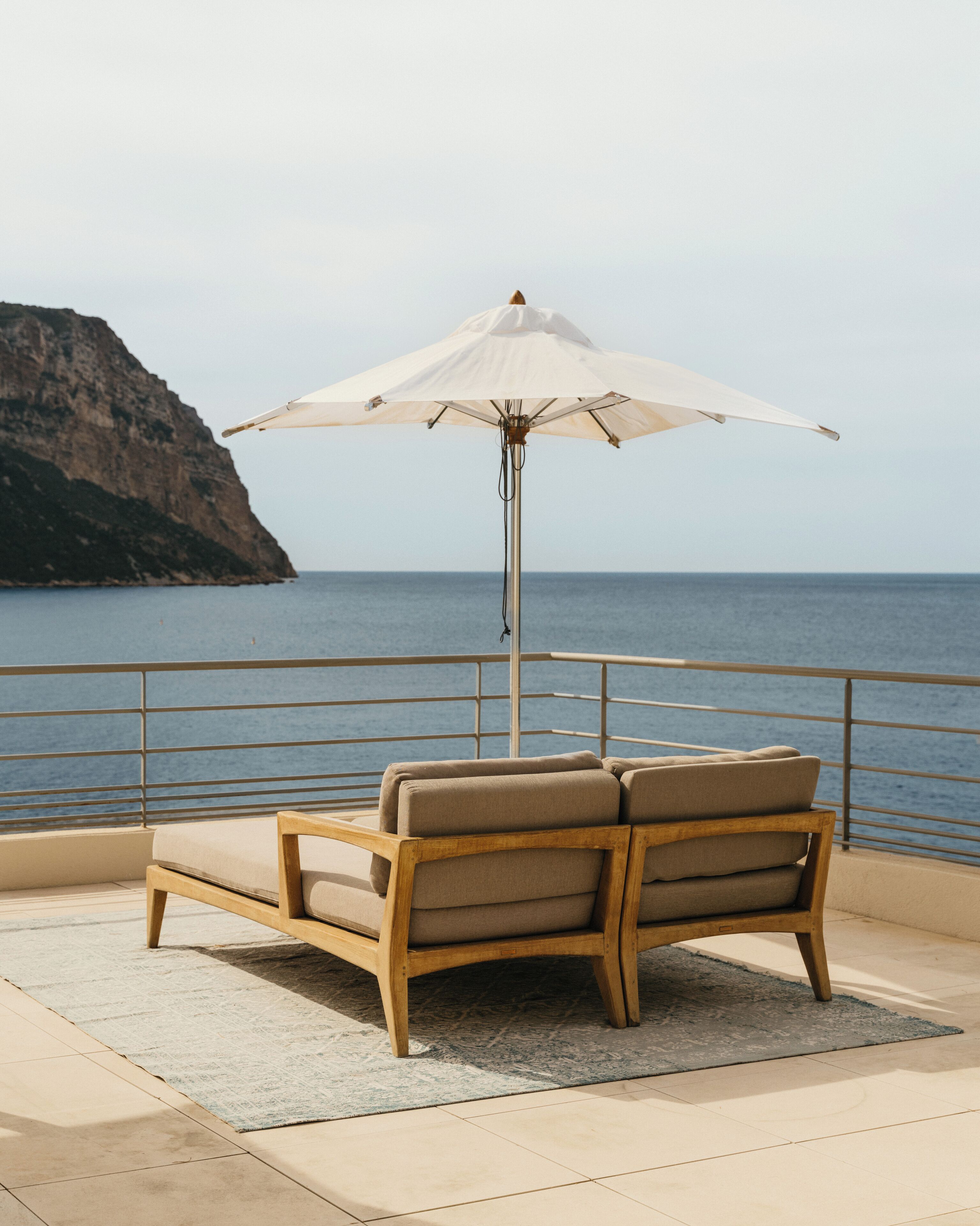 Una silla de madera con un cojín color beige, una sombrilla blanca y vista al mar y un acantilado.