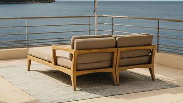 Una silla de madera con un cojín color beige, una sombrilla blanca y vista al mar y un acantilado.