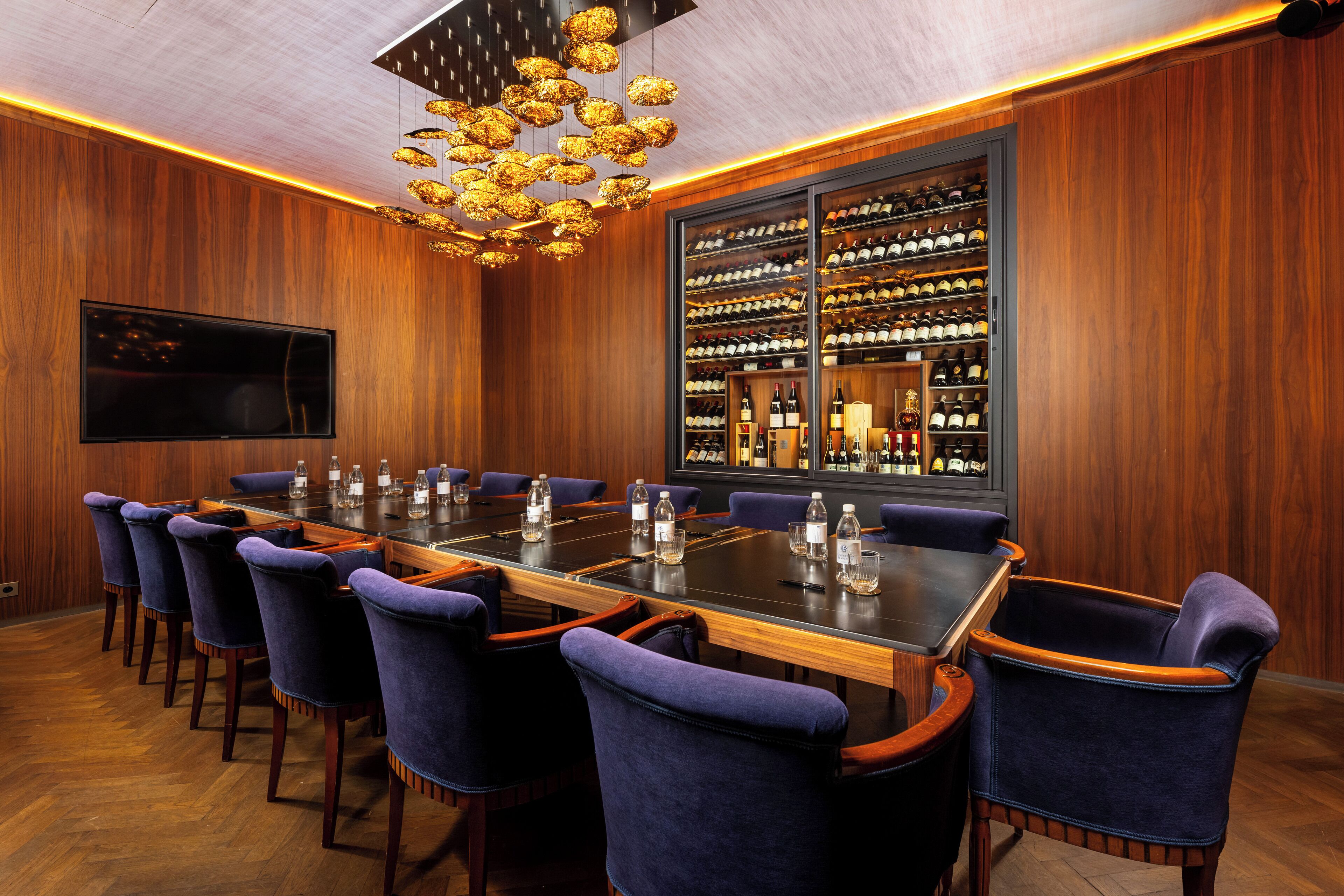 A conference room with a long table, blue upholstered chairs, a wine rack, and a television on the wall.