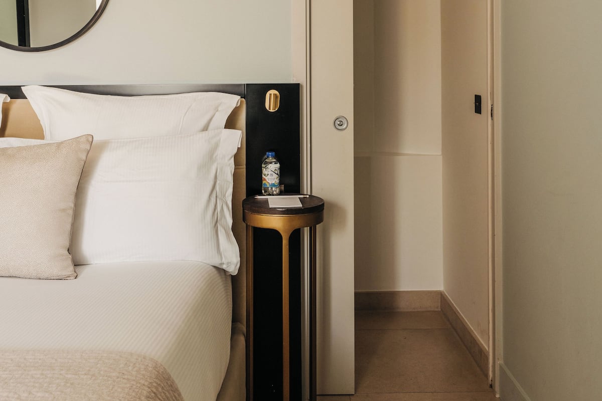 A hotel room with a bed, a round mirror, a small table with a bottle, and a door leading to another room.