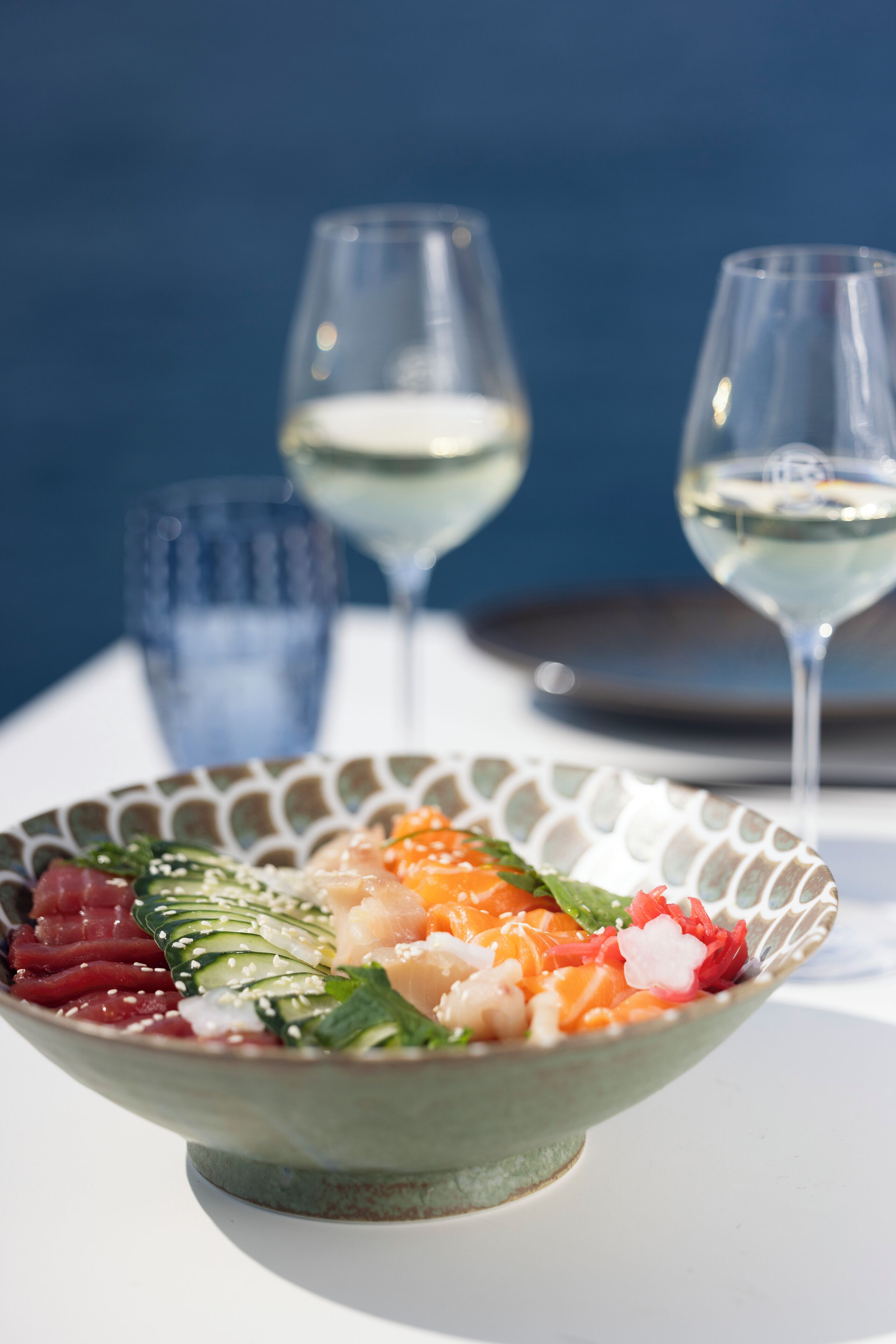 Eine Schüssel Sushi mit verschiedenen Zutaten, darunter Fisch und Gemüse, auf einem weißen Tisch. Im Hintergrund sind zwei Gläser Weißwein zu sehen.