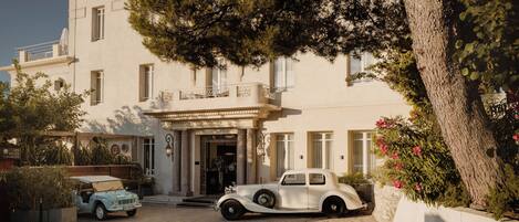 A vintage car parked in front of a building with a stone-paved entrance, columns, and a balcony.