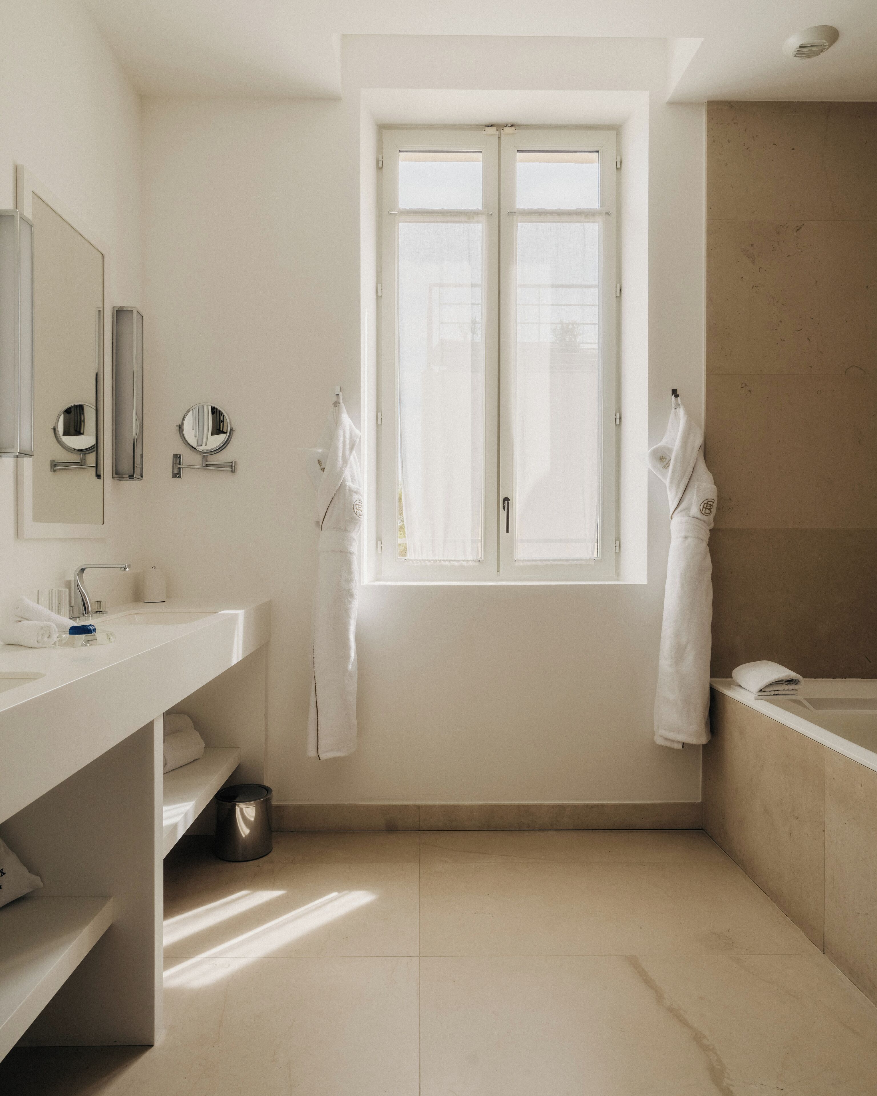 Une salle de bain moderne avec un meuble-lavabo blanc, un grand miroir, deux petits miroirs ronds, une fenêtre avec vue et une baignoire.