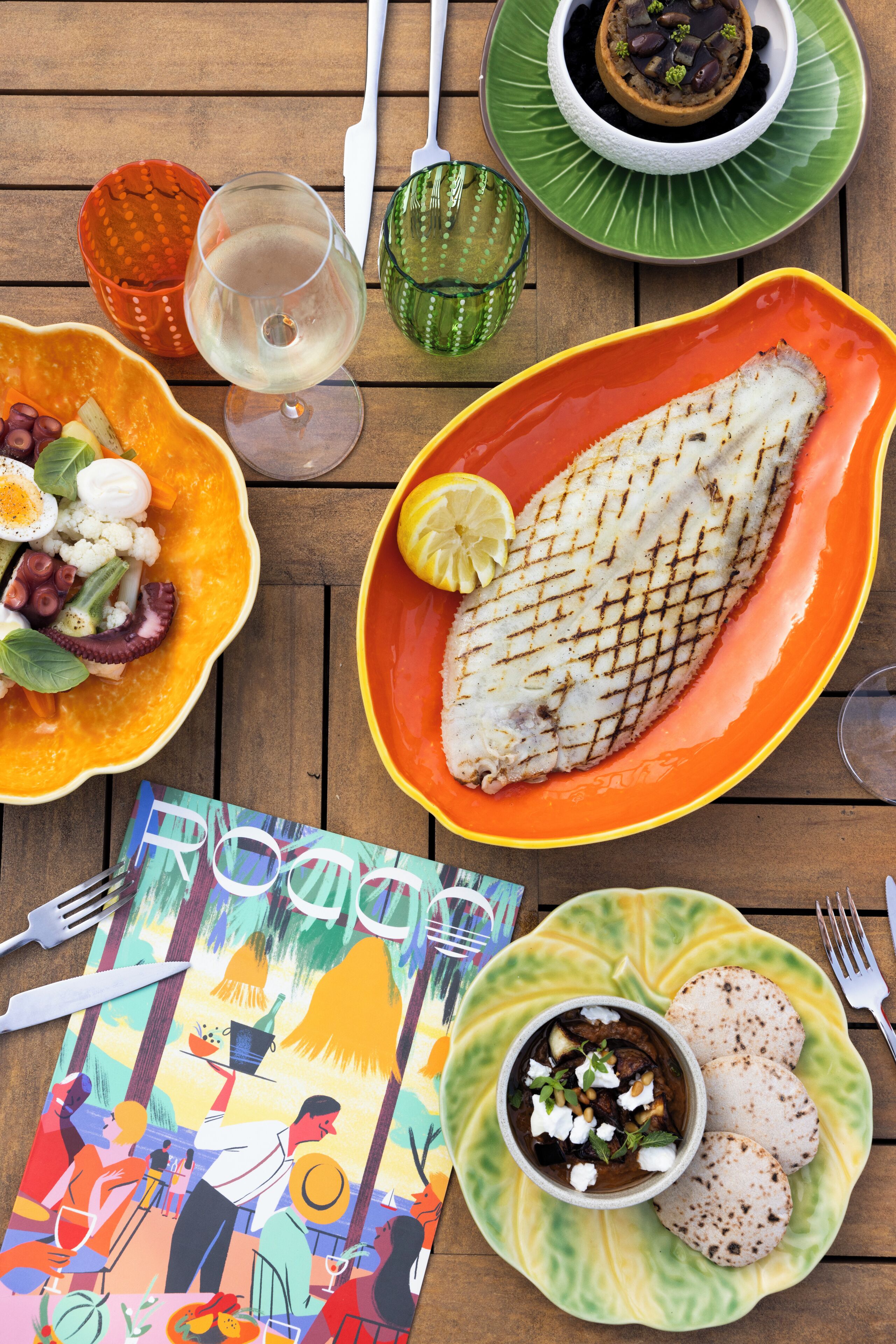 Una mesa con varios platos, incluyendo pescado a la parrilla, un tazón de ensalada y una porción de tortillas.