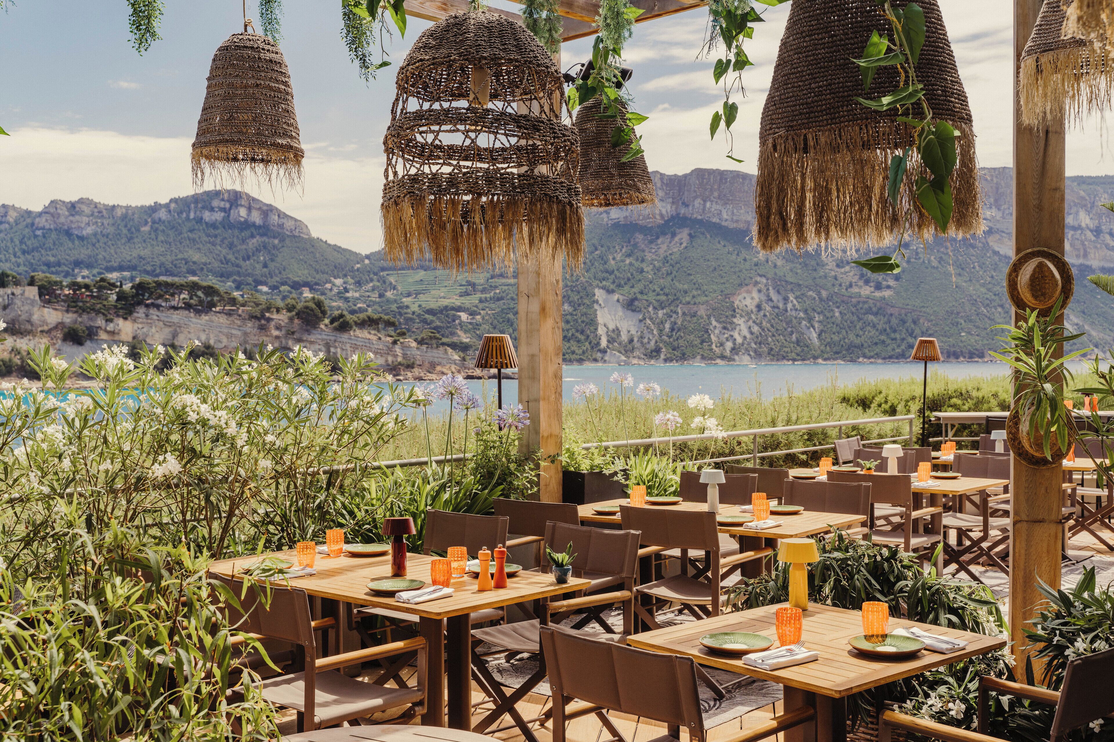 An outdoor dining area with tables and chairs, surrounded by greenery and overlooking a scenic landscape with mountains and a body of water.