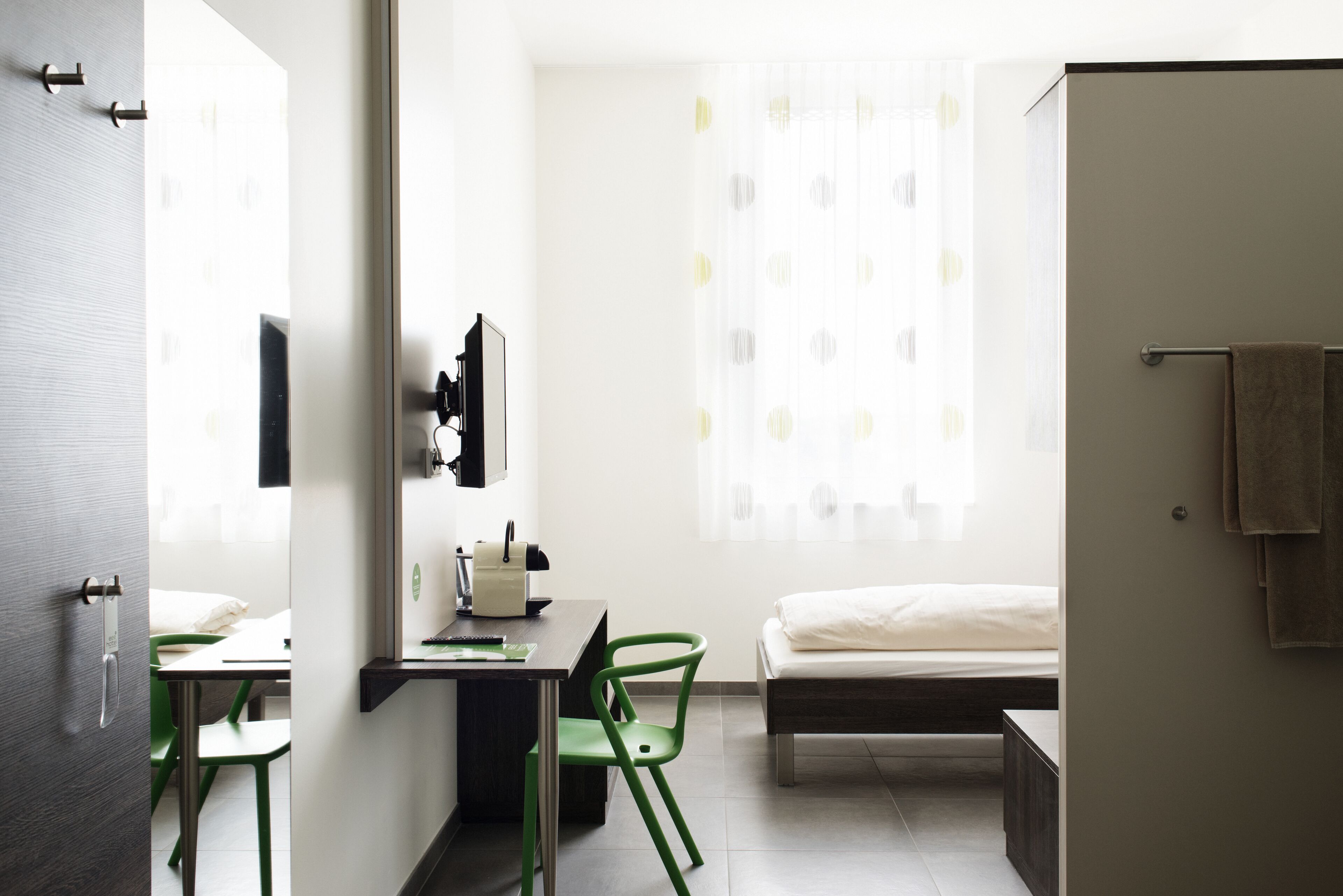 Ein modernes Hotelzimmer mit einem Bett, einem Schreibtisch mit Stuhl, einem Spiegel und einem Vorhangfenster.