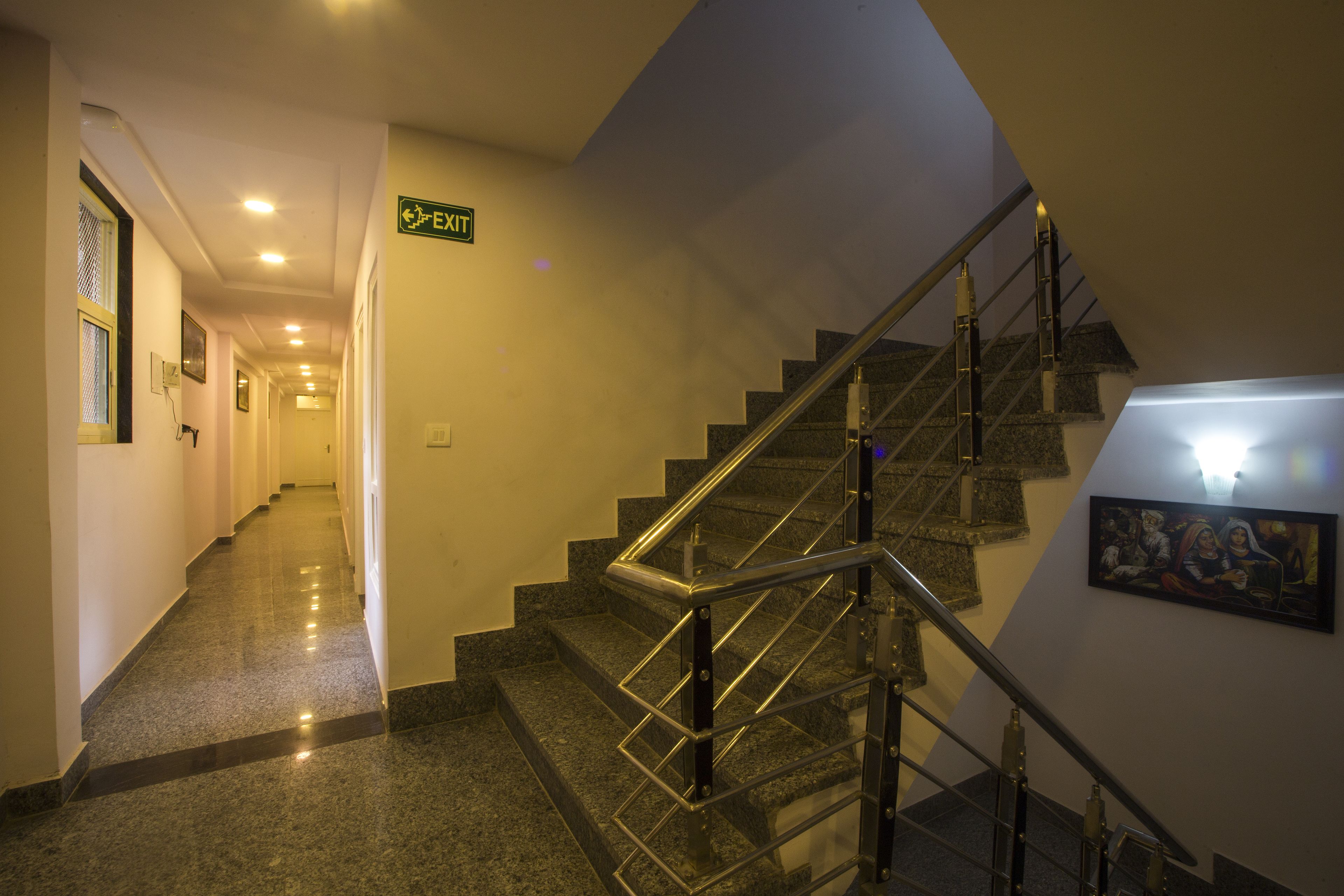 A hotel corridor with a staircase, exit sign, and a framed artwork on the wall.