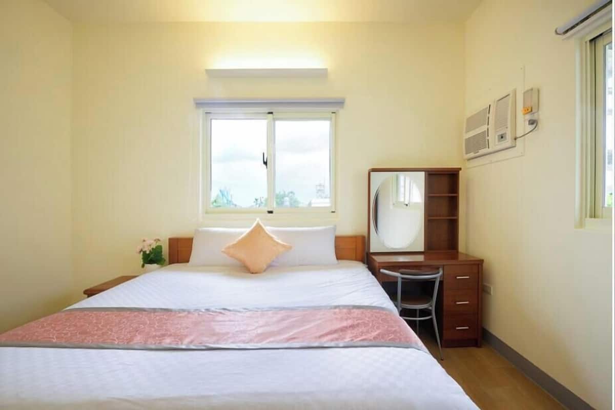 A hotel room with a bed, a desk, a chair, a window, and an air conditioning unit.