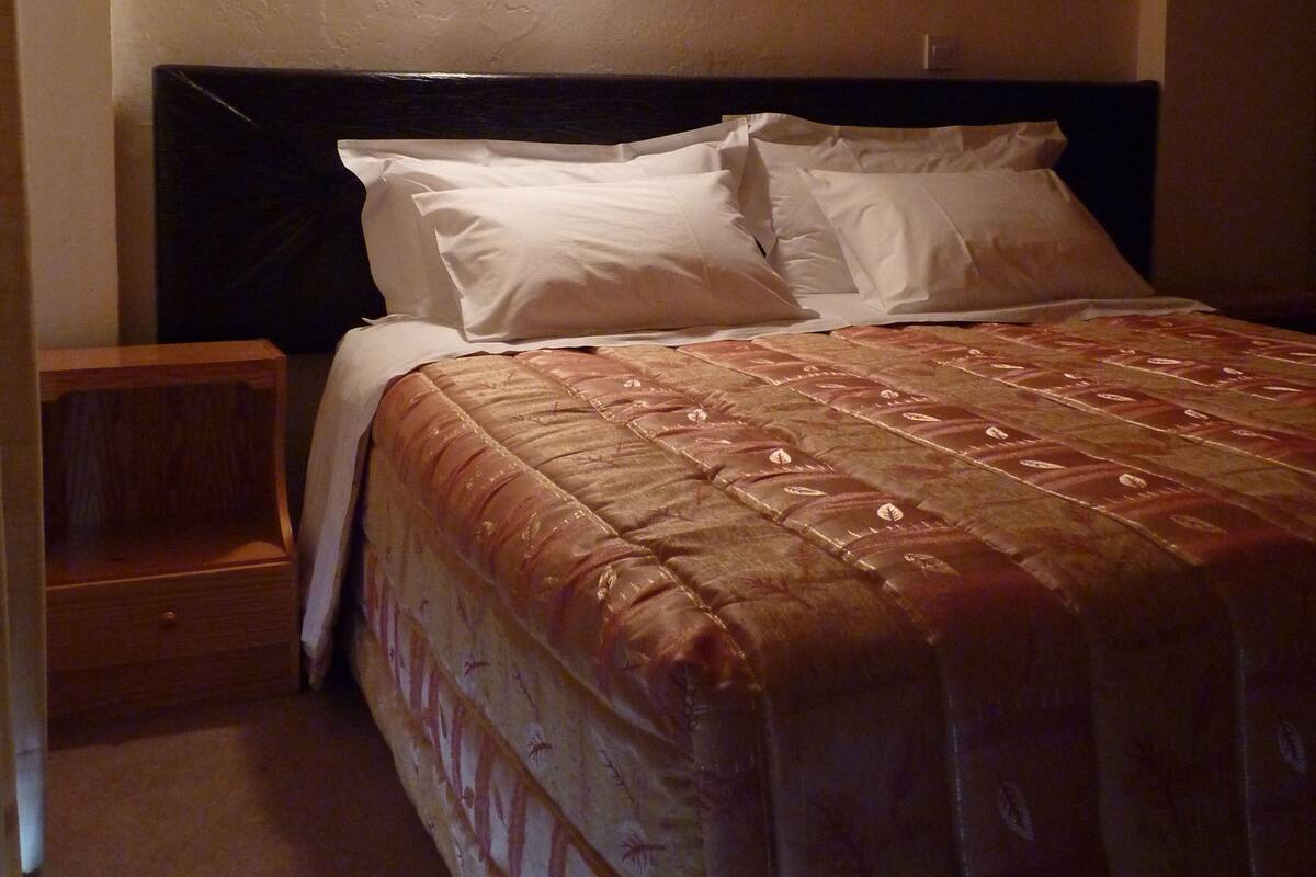 Una cama bien hecha con cabecera, mesita de noche y lámpara de pared.