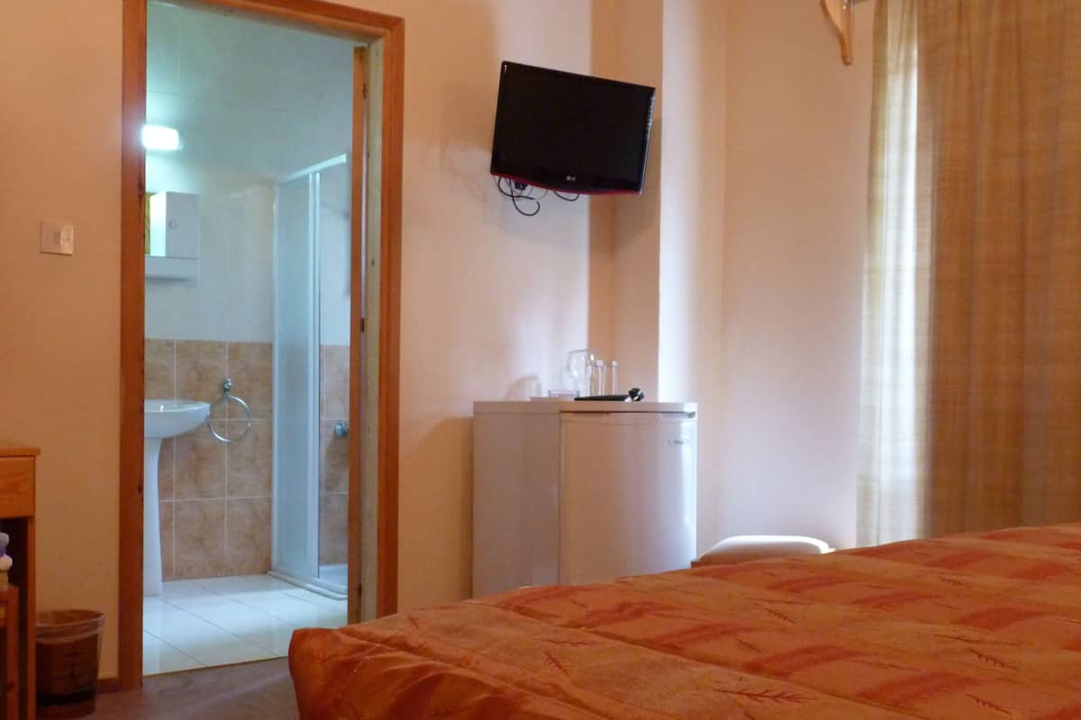 Habitación de hotel con cama, televisor montado en la pared, una mesita y zona de ducha visible a través de una puerta abierta.