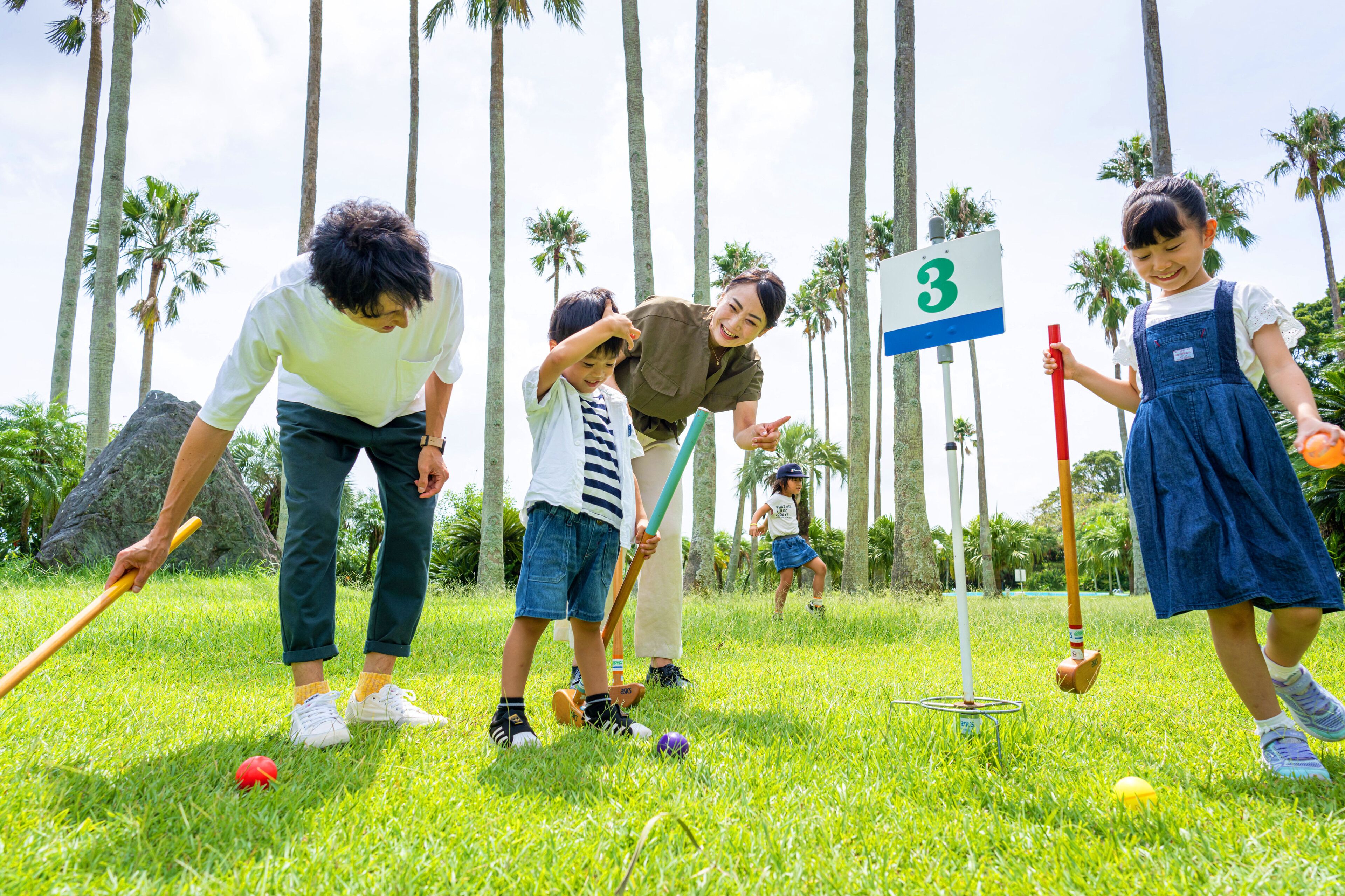 A group of people playing croquet on a grassy field with palm trees in the background.