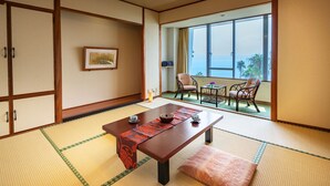 A traditional Japanese room with tatami flooring, a low table with a red patterned table runner, and a view of the ocean through large windows. - Ibusuki Phoenix Hotel (Ibusuki)