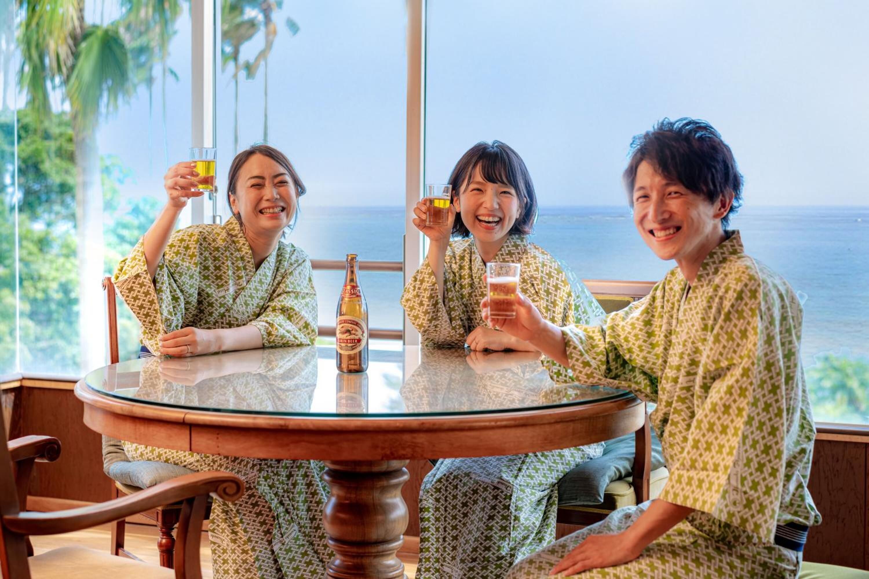 Three people in patterned robes sitting at a round glass table with drinks, overlooking a view of the sea.