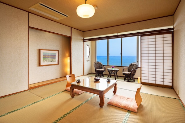 A traditional Japanese room with tatami flooring, a low table, two chairs, and a view of the sea through shoji screens.