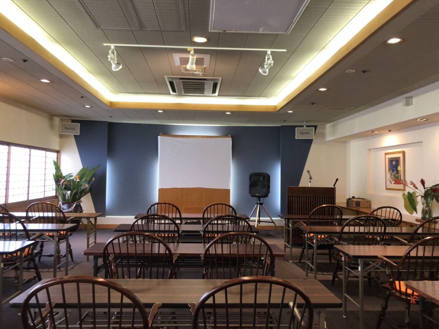 Un salón de conferencias con mesas y sillas, una pantalla de proyector y un micrófono en un atril.