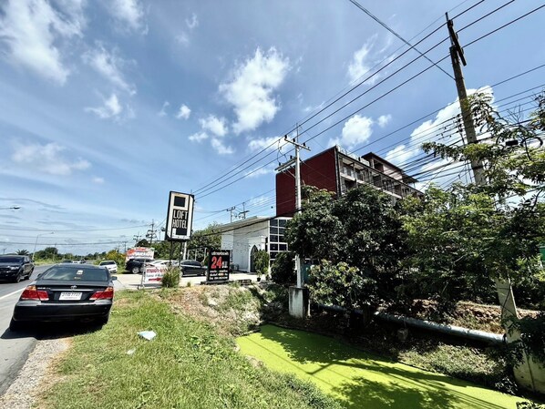 A building with a sign for a hotel, a parking lot, and a road with cars. - PJ Loft Hotel (Lop Buri)