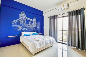 A hotel room with a large bed, a blue wall with a bridge illustration, a balcony with curtains, and a window. - PJ Loft Hotel (Lop Buri)