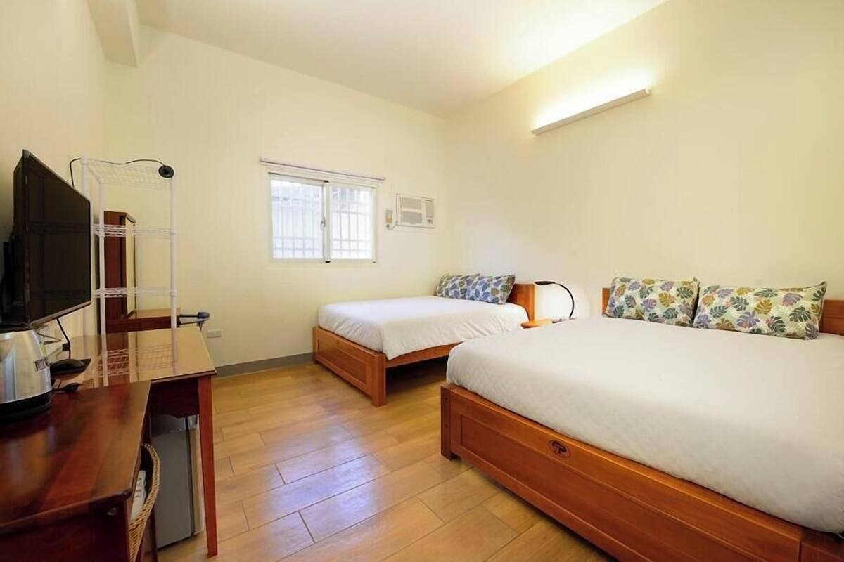 A bedroom with two single beds, a wooden desk with a television, and a window with blinds.
