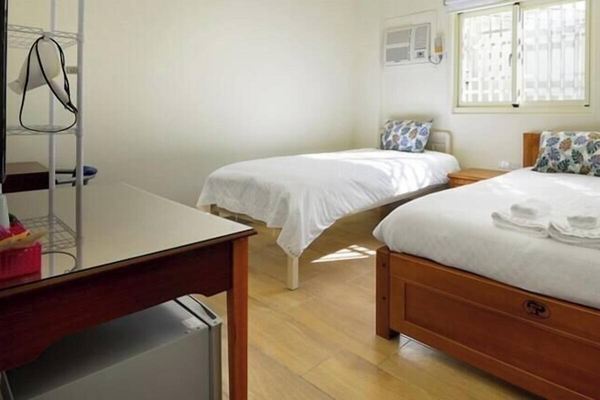 A hotel room with two single beds, a wooden desk, a chair, a small table, and a window with blinds.