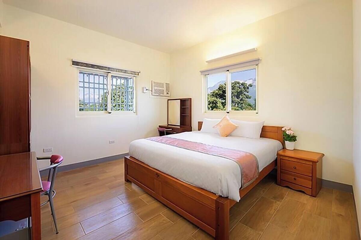 A bedroom with a wooden bed, two bedside tables, a nightstand, a chair, and two windows with blinds.