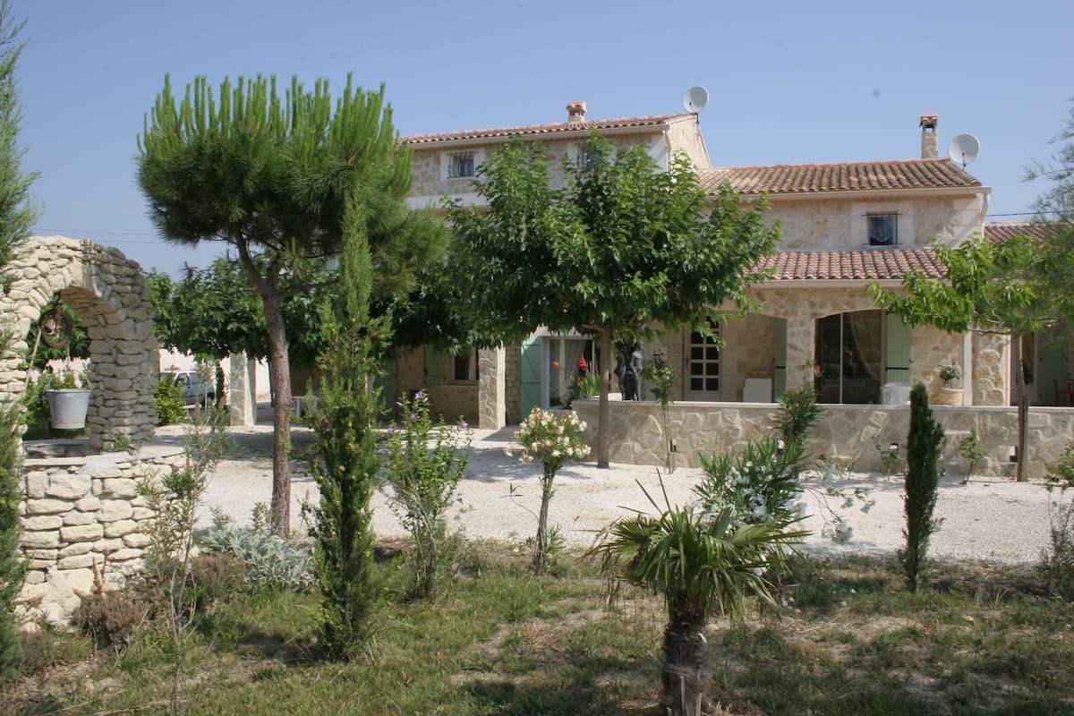 A stone house with a tiled roof, surrounded by a gravel courtyard and various plants.