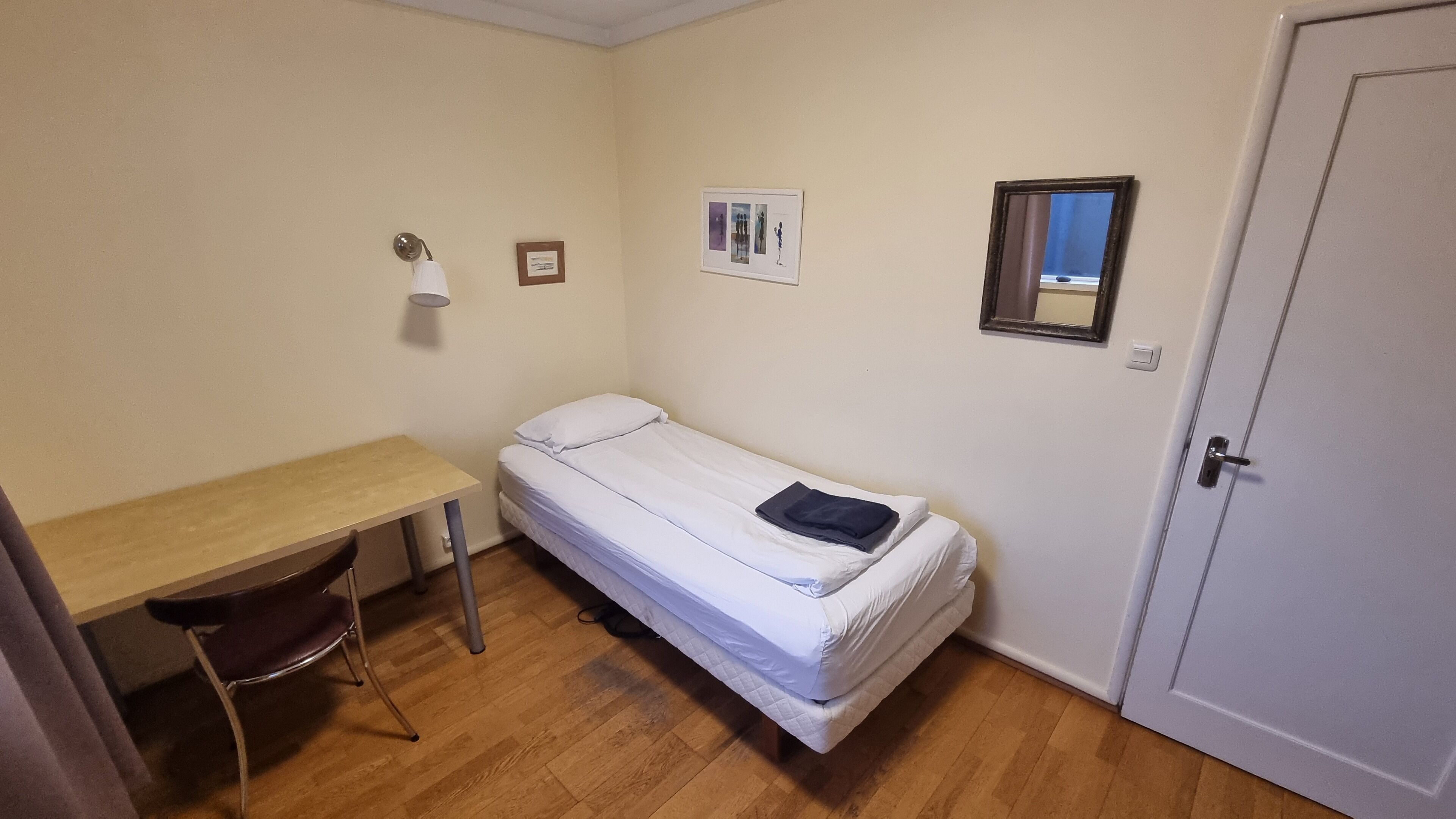 Una habitación pequeña con cama individual, un escritorio de madera y una silla, una mesita de noche con lámpara, y una puerta.