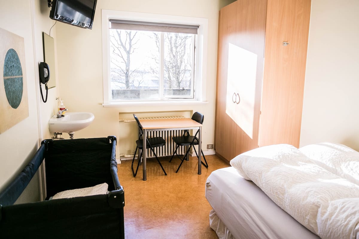Une chambre avec un lit, un bureau avec deux chaises, un radiateur, une fenêtre donnant sur des arbres et une valise noire posée sur le sol.