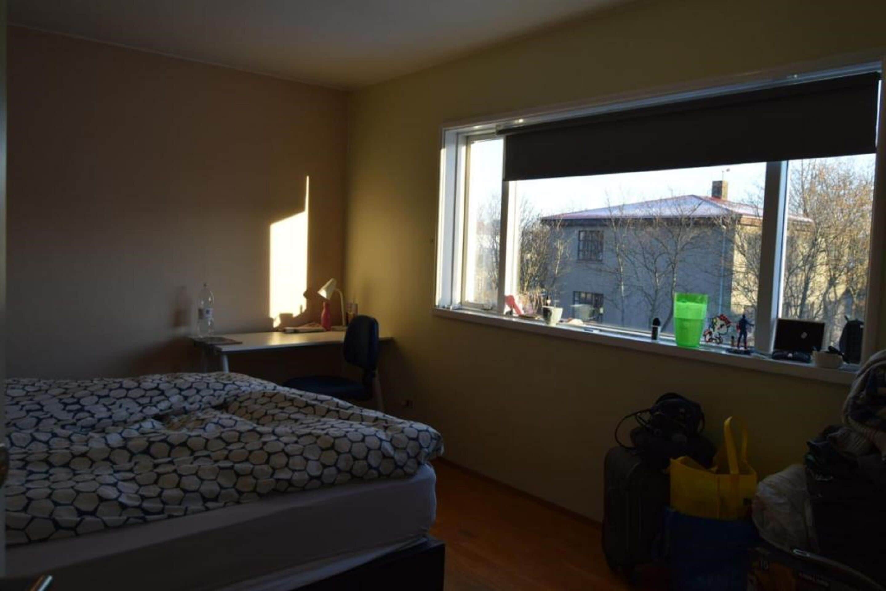 Un dormitorio con cama, escritorio y ventana con vista a una casa.