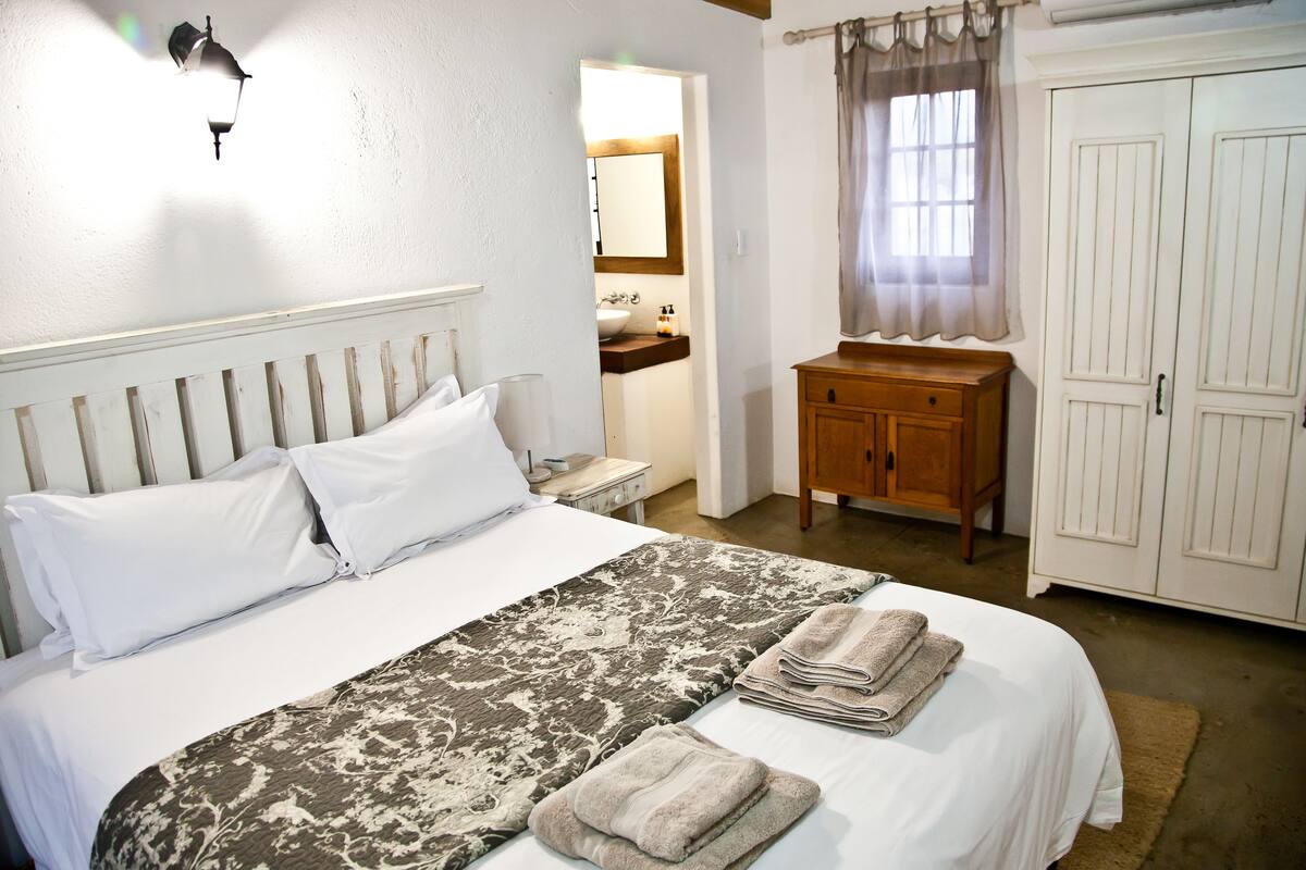 A neatly made bed with white linens and a patterned throw, a wooden nightstand with a lamp, a wooden dresser, and a bathroom visible through an open door.