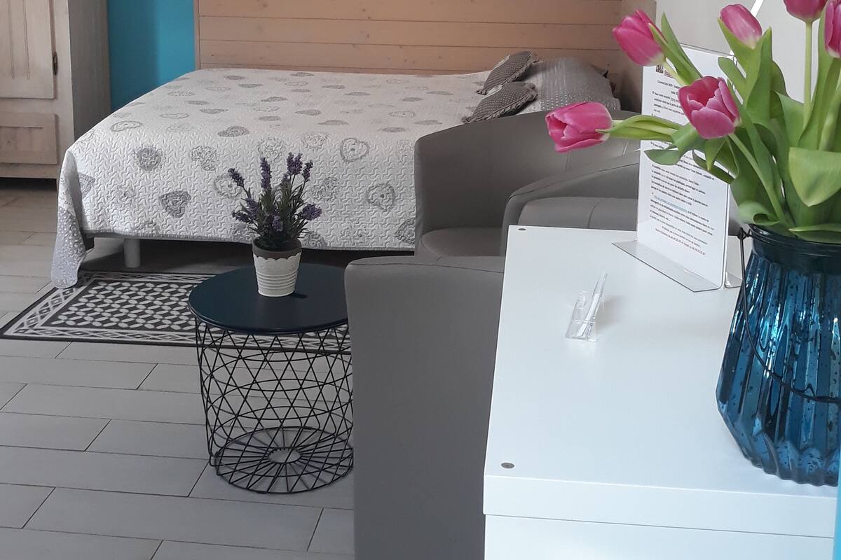 A bedroom with a bed, a desk, a chair, a small table with a plant, and a vase with flowers.