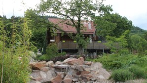 Eine rustikale Holzhütte mit rotem Ziegeldach, umgeben von üppiger Vegetation und einem kleinen Wasserfall.