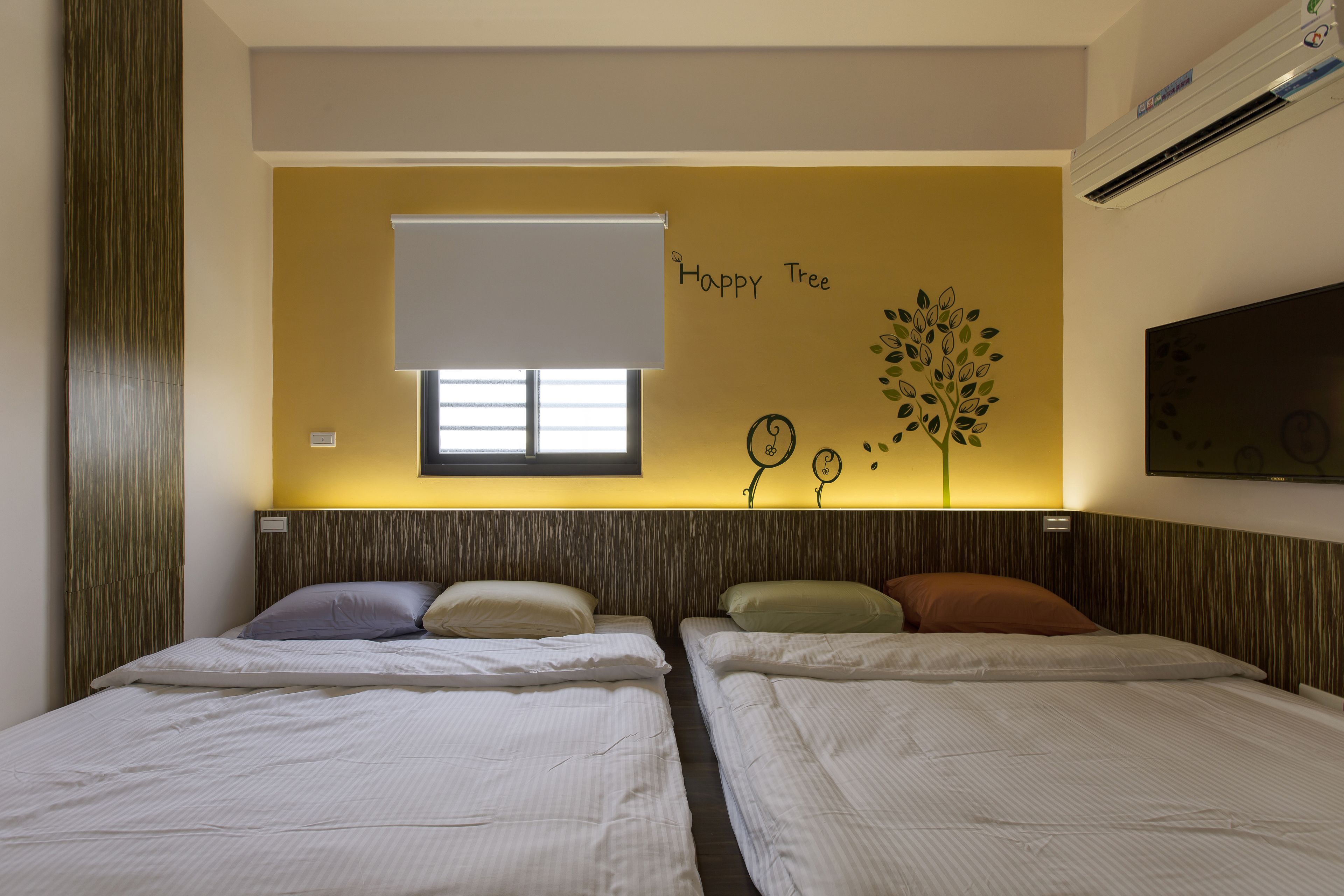 A hotel room with two beds, a window with a blind, a wall with a 'Happy Tree' decoration, and an air conditioning unit.