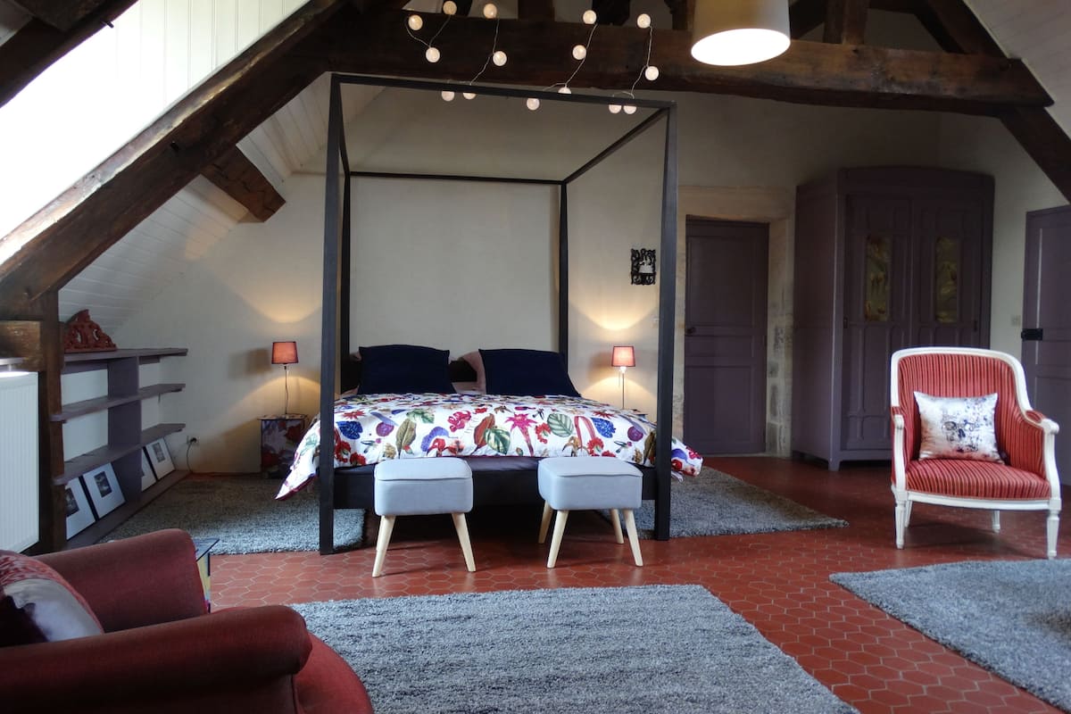 Een hemelbed met een bloemenprint, een rode fauteuil, een grijze poef en een vloer van rode bakstenen.