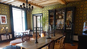 Een eetruimte met een houten tafel, stoelen en een kroonluchter. Er is een groot raam met gordijnen en een potplant.