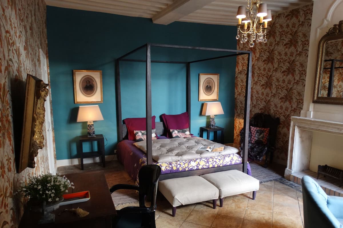 Een hemelbed met een baldakijn, twee nachtkastjes met lampen, een stoel, een bankje en een bureau met een bloevas.