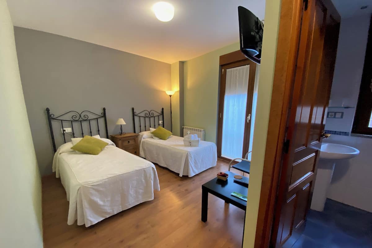 Quarto de hotel com duas camas de solteiro, uma mesinha, televisão e banheiro com pia.