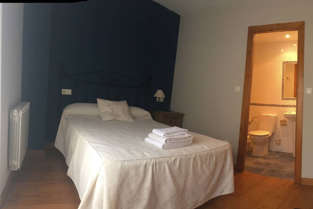 Quarto com cama, mesa de cabeceira, abajur e vista para o banheiro.