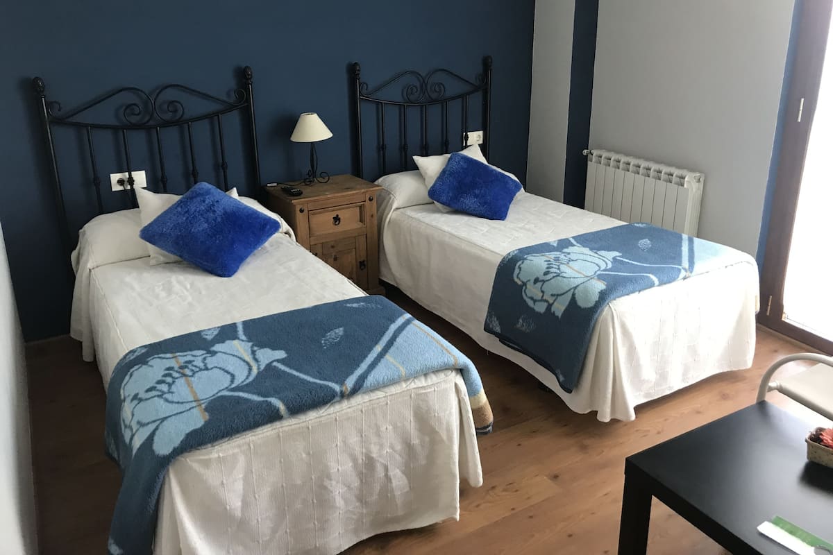 Quarto com duas camas, cada uma com colcha azul florida e travesseiro azul. Há uma mesinha de cabeceira de madeira com um abajur entre as camas.