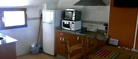 Una cocina con microondas, estufa y una campana extractora grande. Hay una escoba apoyada en la pared y piso de baldosas.