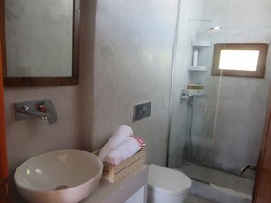 A bathroom with a large basin, a toilet, a glass shower enclosure, and a wooden shelf with a towel.