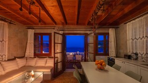 Un cuarto acogedor con techo de madera, una mesa de centro de vidrio con velas, una mesa de comedor con frutero y vistas al mar a través de puertas abiertas.