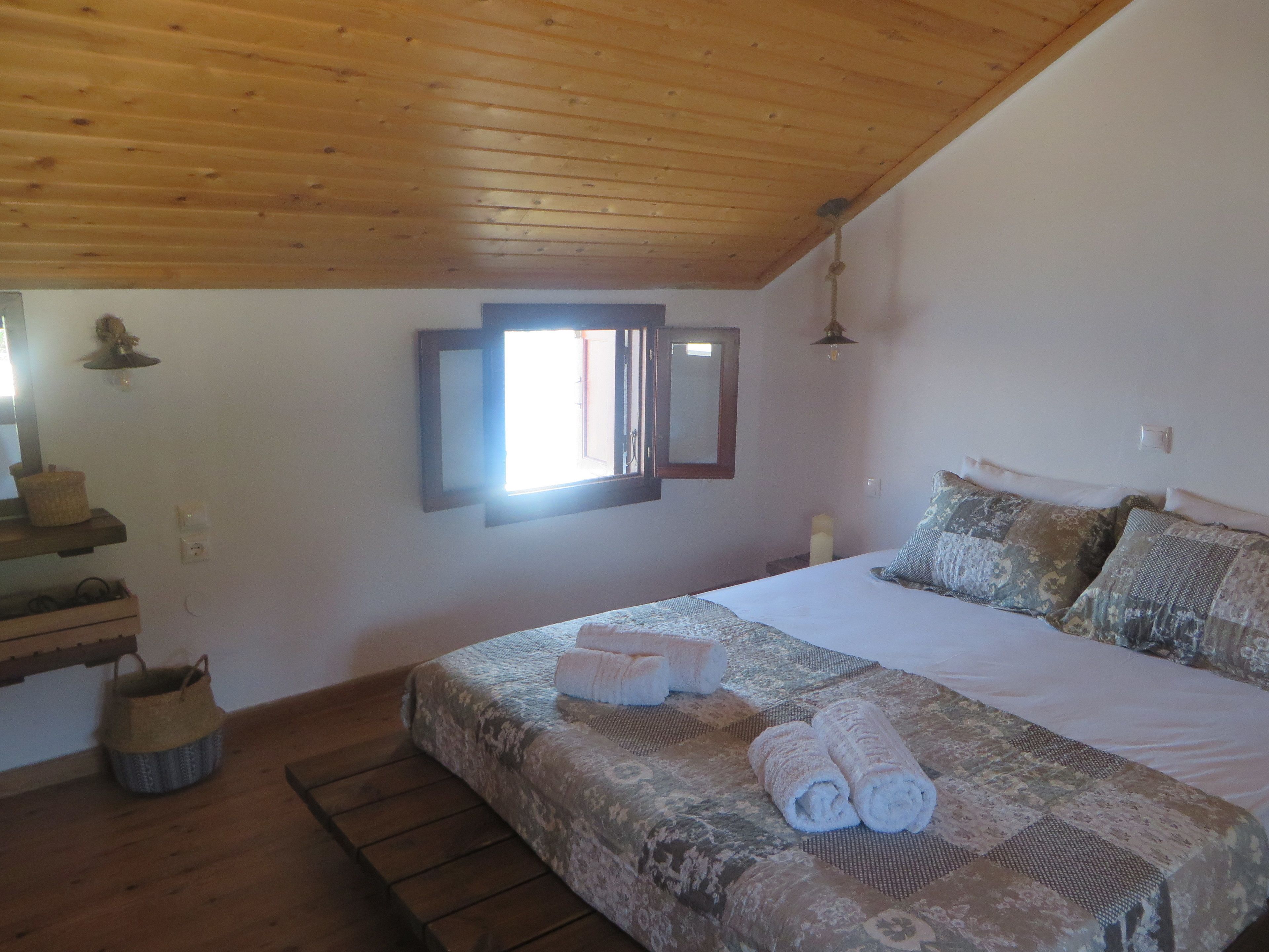 Une chambre à coucher avec un plafond en bois, un lit avec une literie à motifs, une lampe de chevet et une fenêtre donnant sur un beau paysage.