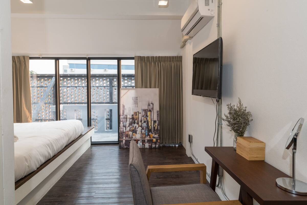 A hotel room with a bed, a desk, a chair, a TV, and a large window with a view of buildings.